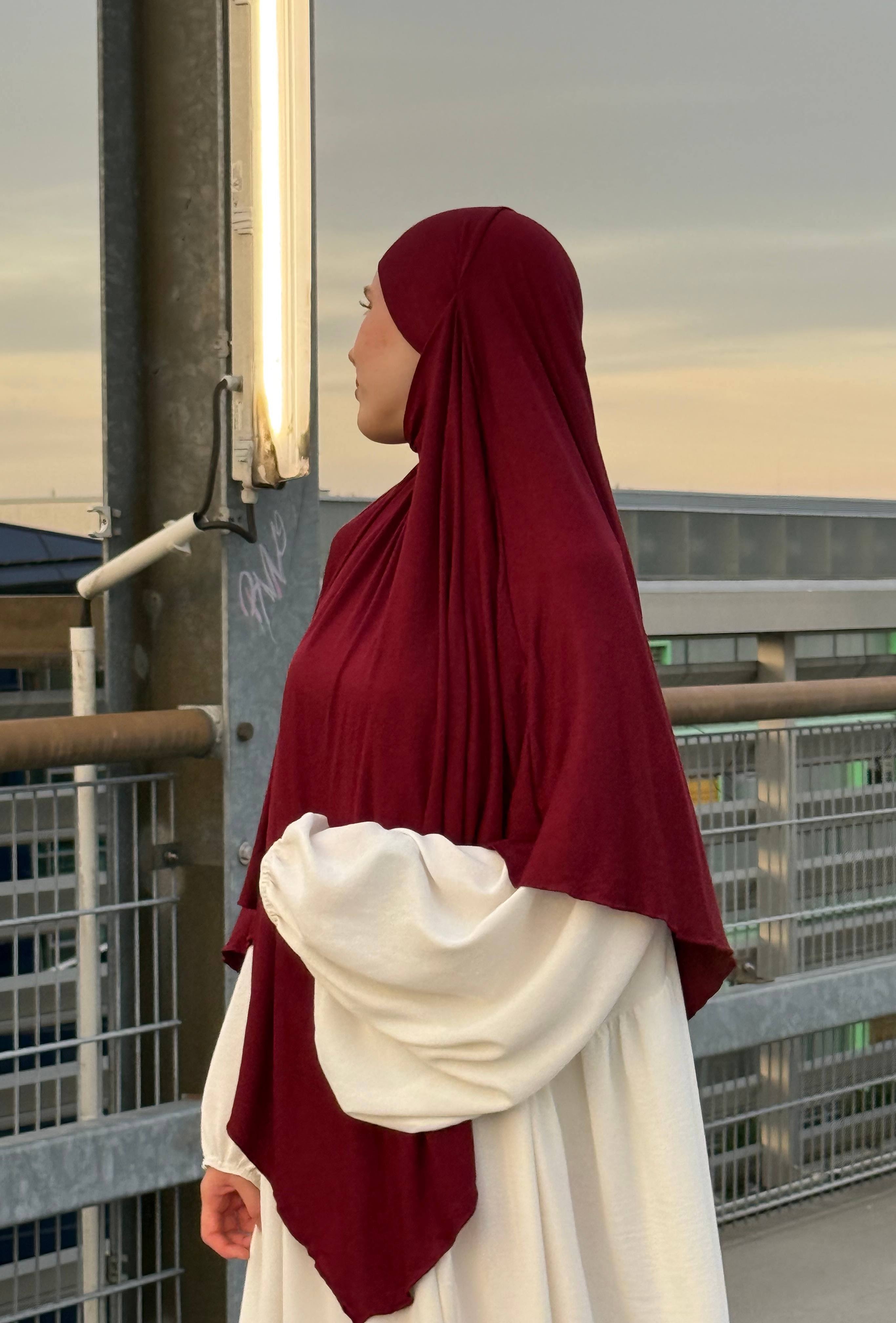 Jersey Khimar in bordeauxrot aus Viskose-Elastan, seitliche Ansicht einer Person vor Geländer auf einem Parkdeck.