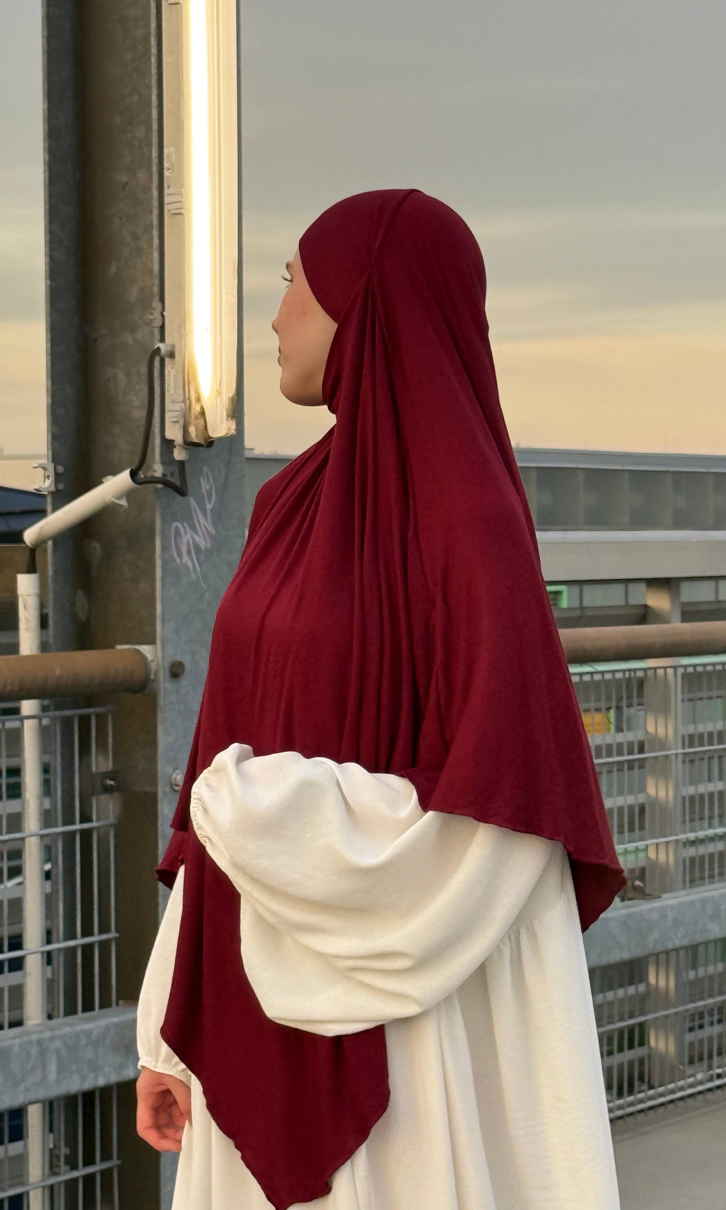Jersey Khimar in bordeauxrot aus Viskose-Elastan, seitliche Ansicht einer Person vor Geländer auf einem Parkdeck.