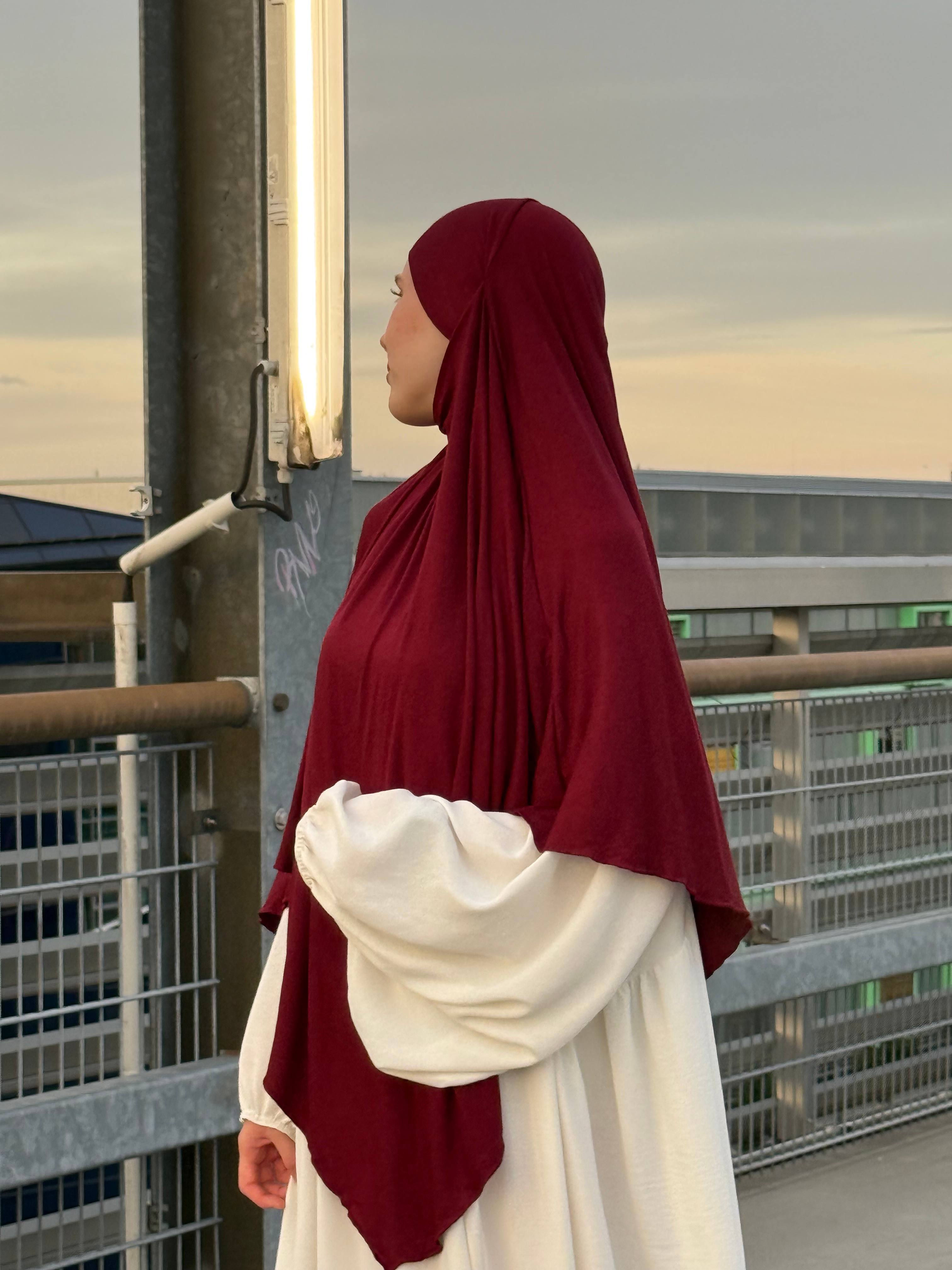 Jersey Khimar in bordeauxrot aus Viskose-Elastan, seitliche Ansicht einer Person vor Geländer auf einem Parkdeck.