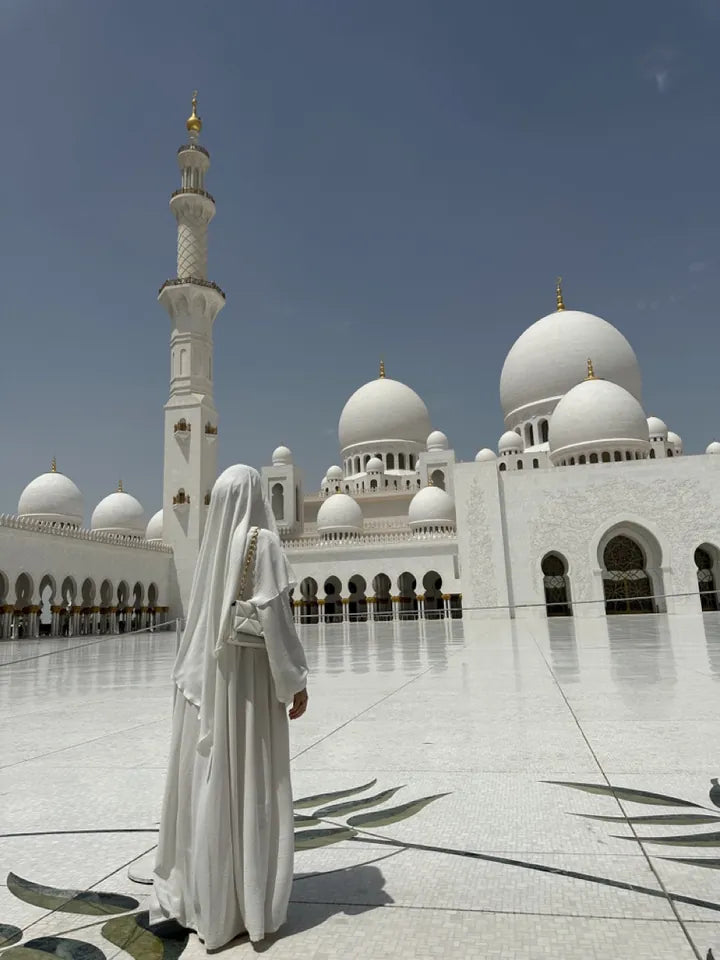 Eine Frau in weißer Abaya und Hijab steht mit dem Rücken zur Kamera vor der prachtvollen, weißen Sheikh Zayed Moschee.