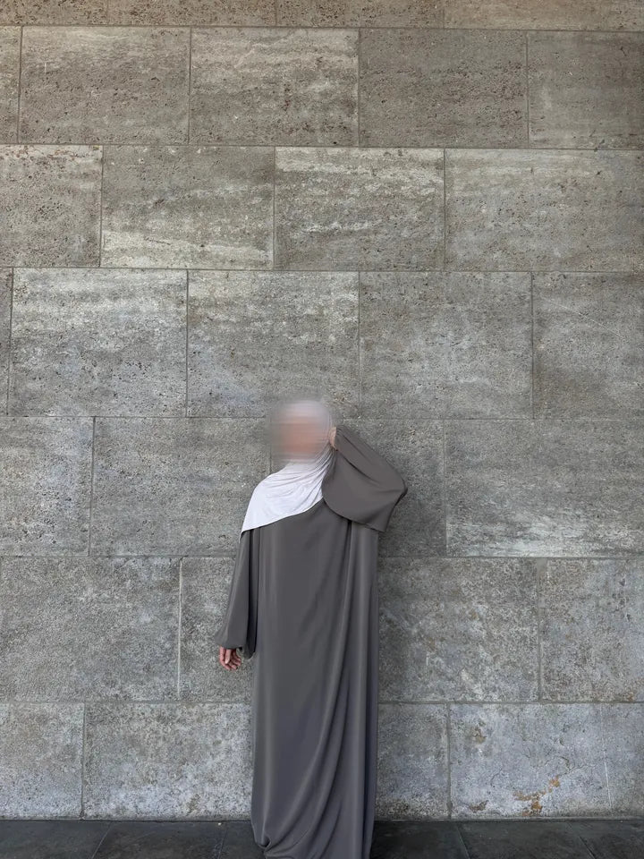 Eine Person trägt eine lange, taupefarbene Abaya und einen hellen Hijab. Eine Hand ist zum Kopf gehoben, um den Hijab anzupassen, wodurch der weite Ärmel zur Geltung kommt.