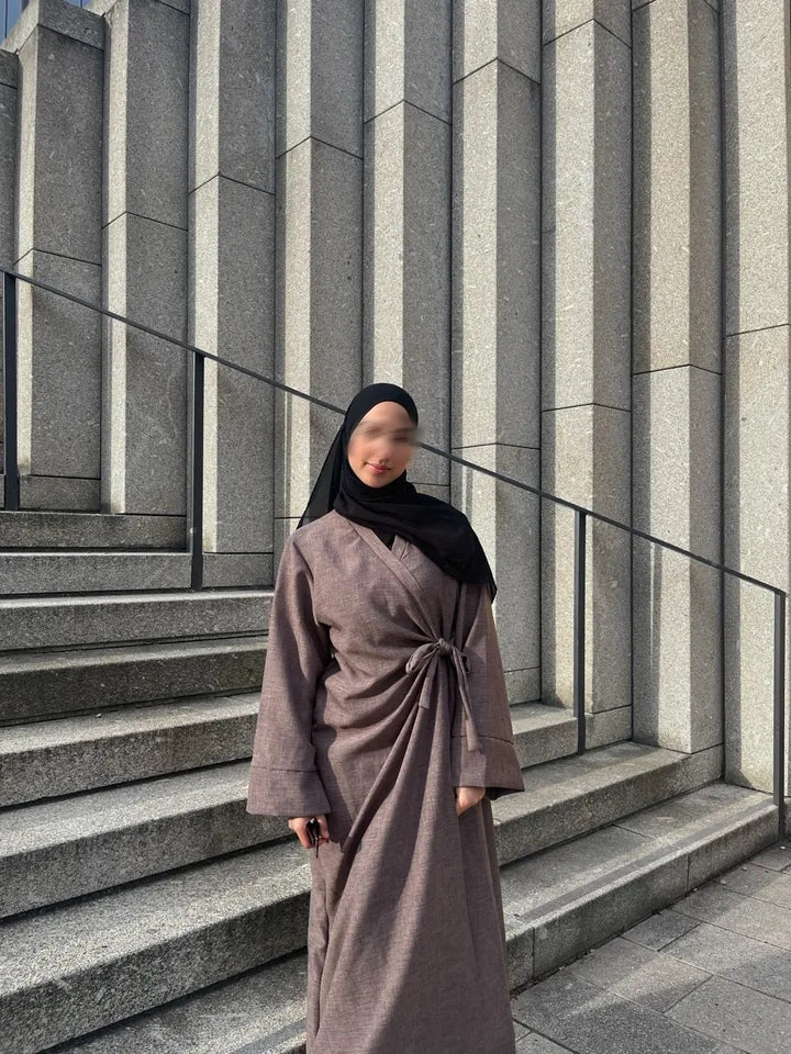 Eine Frau trägt eine lange, braune Wickel-Abaya mit seitlichem Bindegürtel und einen schwarzen Hijab, während sie auf einer Steintreppe steht.