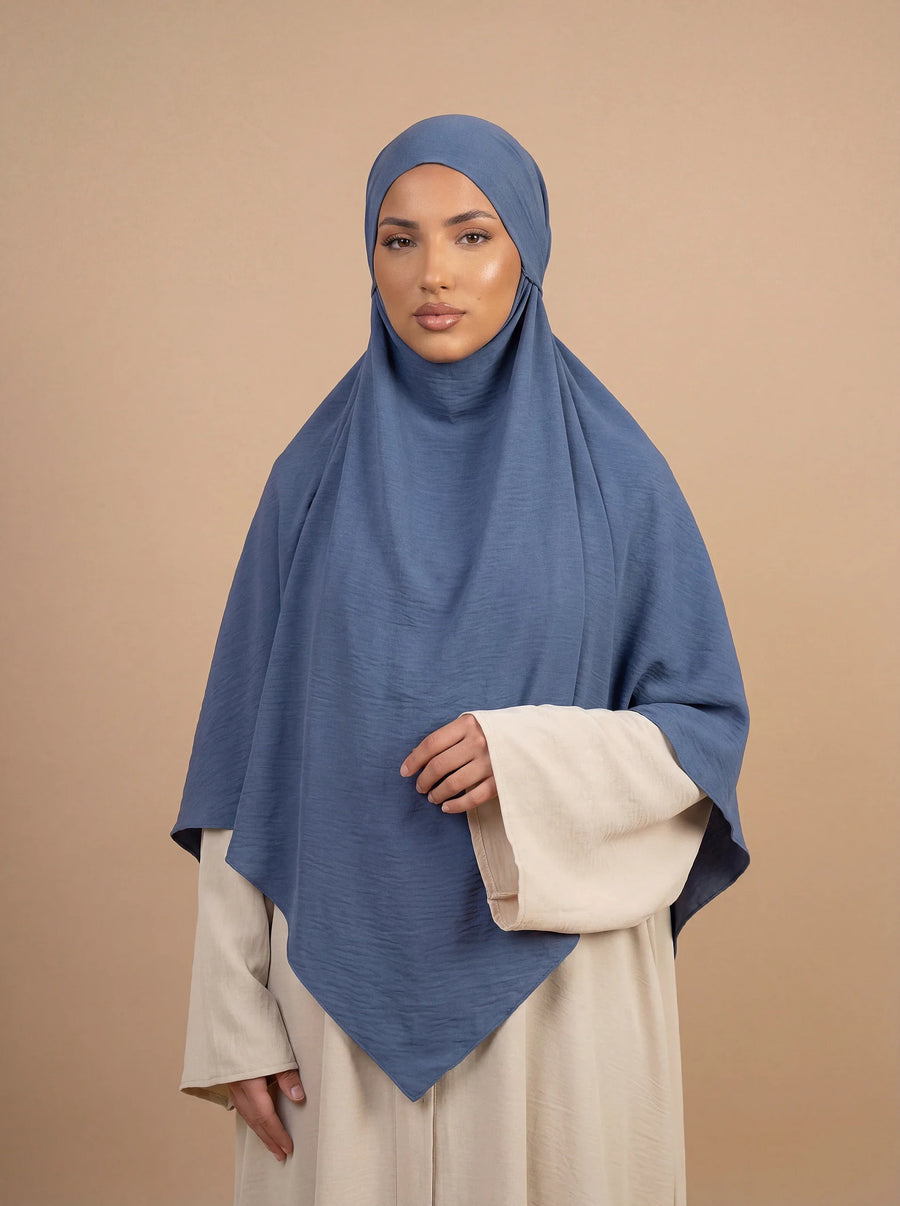 Unser langer Khimar in der Farbe Taupe, getragen von einem Model vor einer Wand mit dekorativen Blumen. Die fließende Silhouette des Khimar wird von der Seite gezeigt.