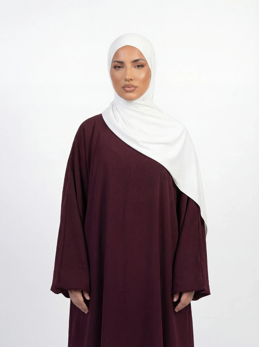 Eine Frau, ganz in Weiß gekleidet, steht auf dem weiten, glänzenden Marmorhof der Sheikh Zayed Moschee und blickt auf die strahlend weißen Kuppeln und das Minarett unter einem hellblauen Himmel.