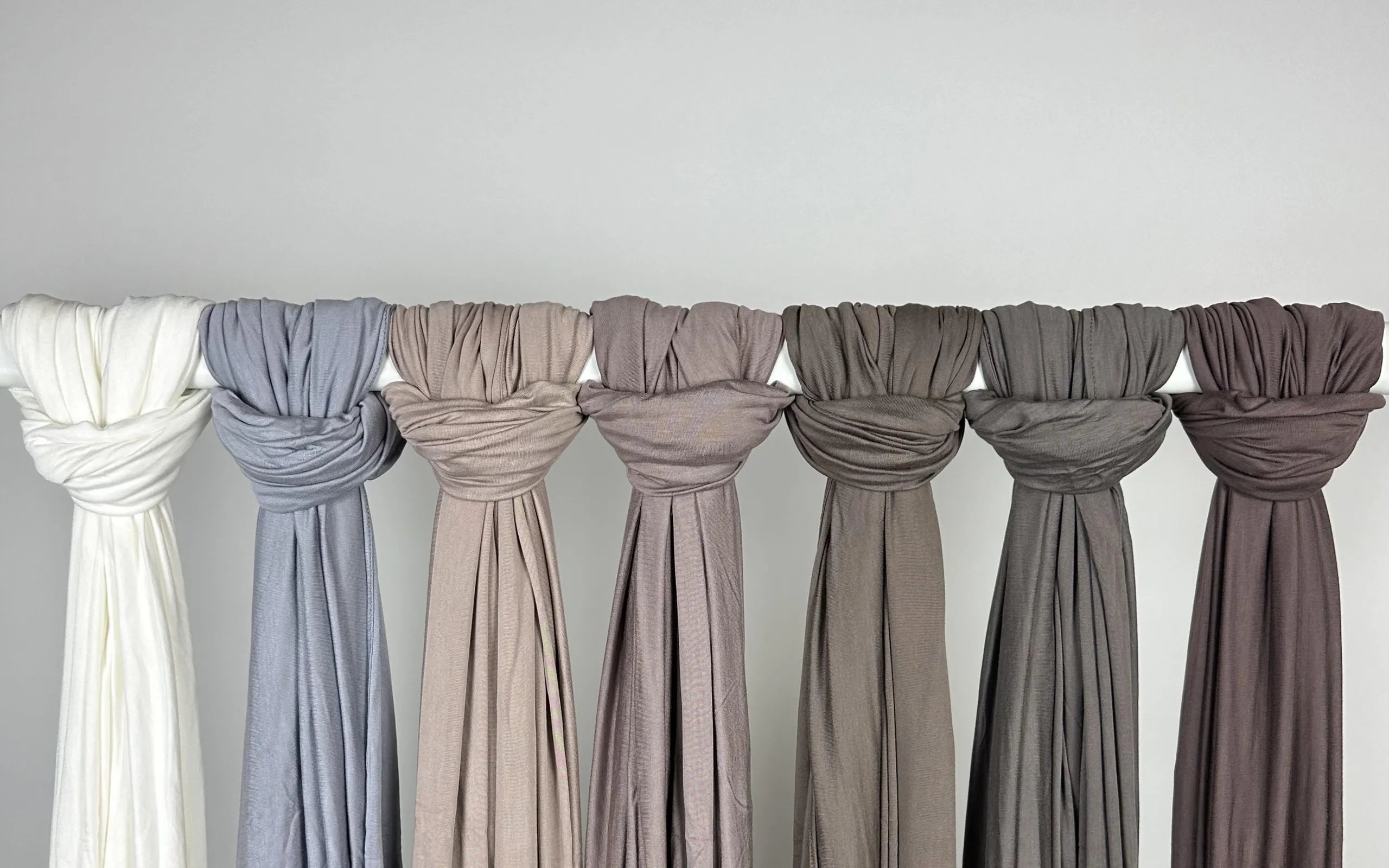 Farbpalette von Jersey-Hijabs in neutralen und erdigen Tönen. Sieben Tücher in Weiß, Blaugrau, Beige, Mauve und verschiedenen Brauntönen hängen an einer Präsentationsstange vor einer grauen Wand.