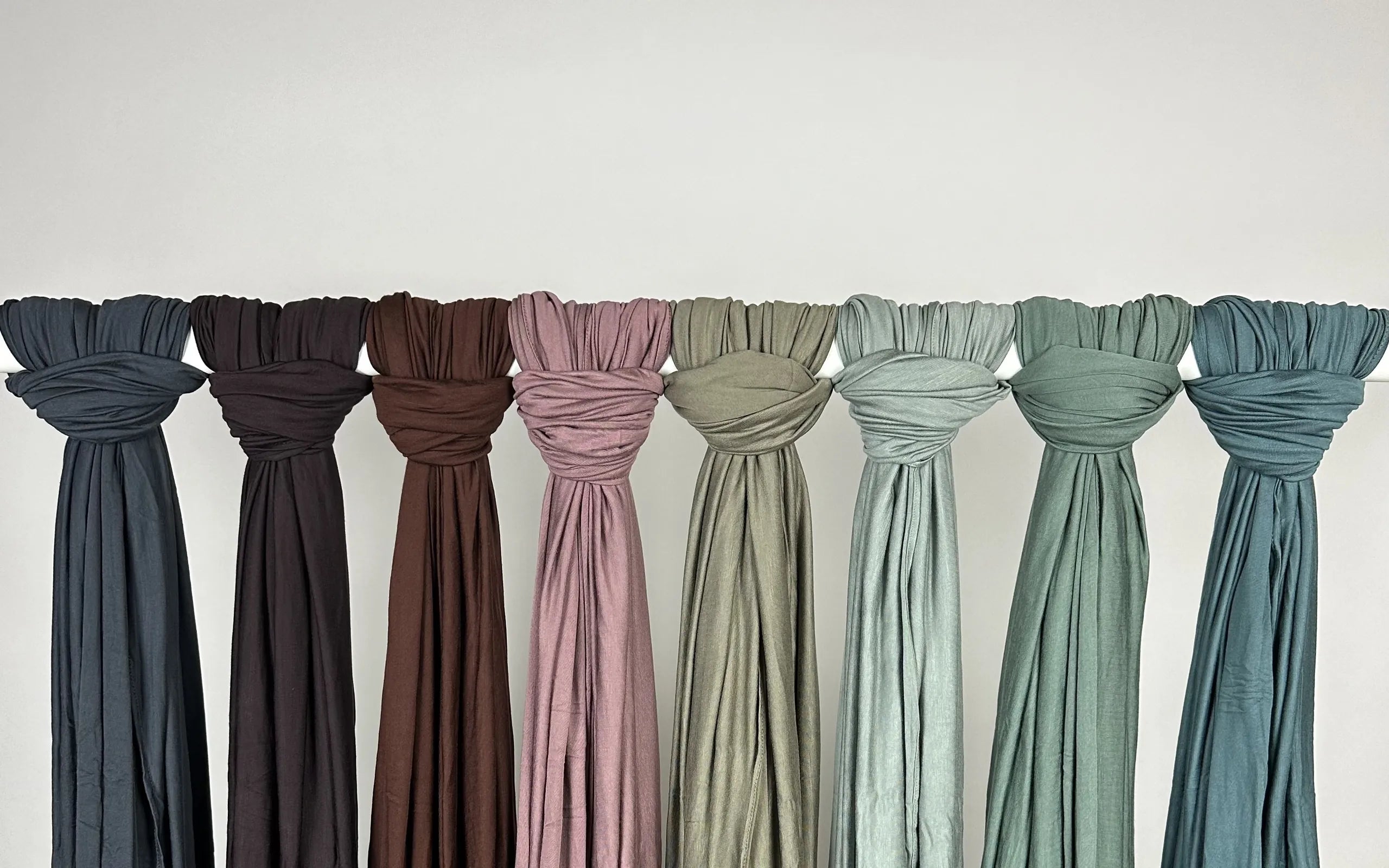 Farbpalette von Jersey-Hijabs in verschiedenen Grün-, Blau-, Braun- und Rosatönen. Die acht Tücher aus weichem Stoff hängen zur Präsentation an einer Stange vor einer grauen Wand.
